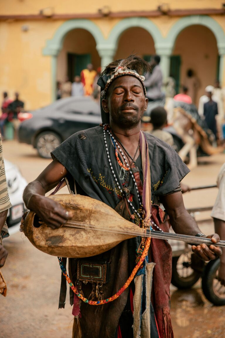 An African musician in traditional attire playing an instrument on a lively street.